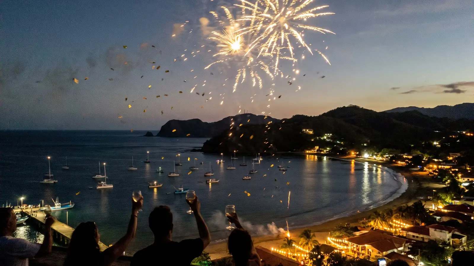 Fireworks over the bay at dusk, with glasses raised in the foreground