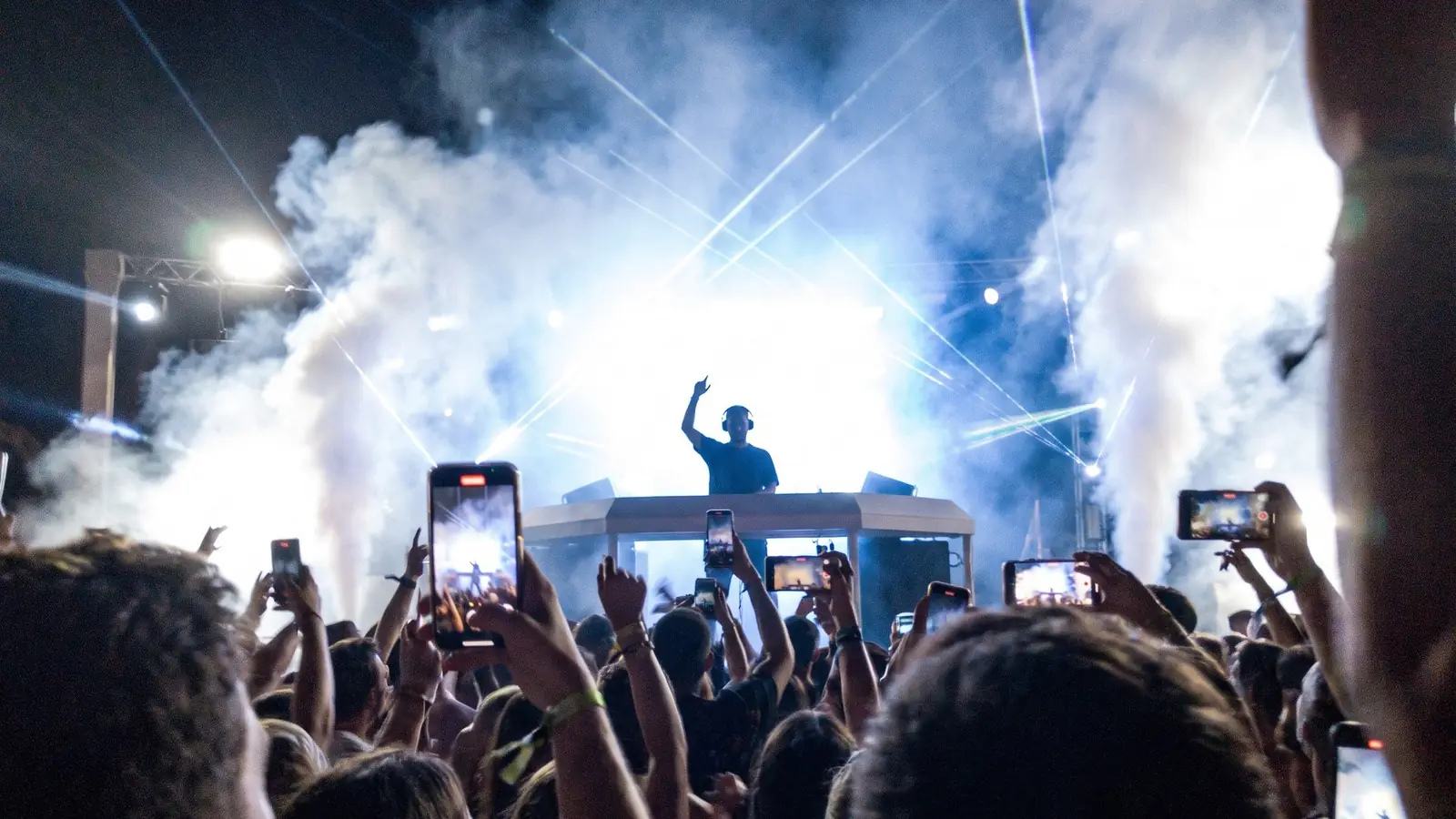 A DJ booth seen from the crowd, with haze, blue lights and phones held up