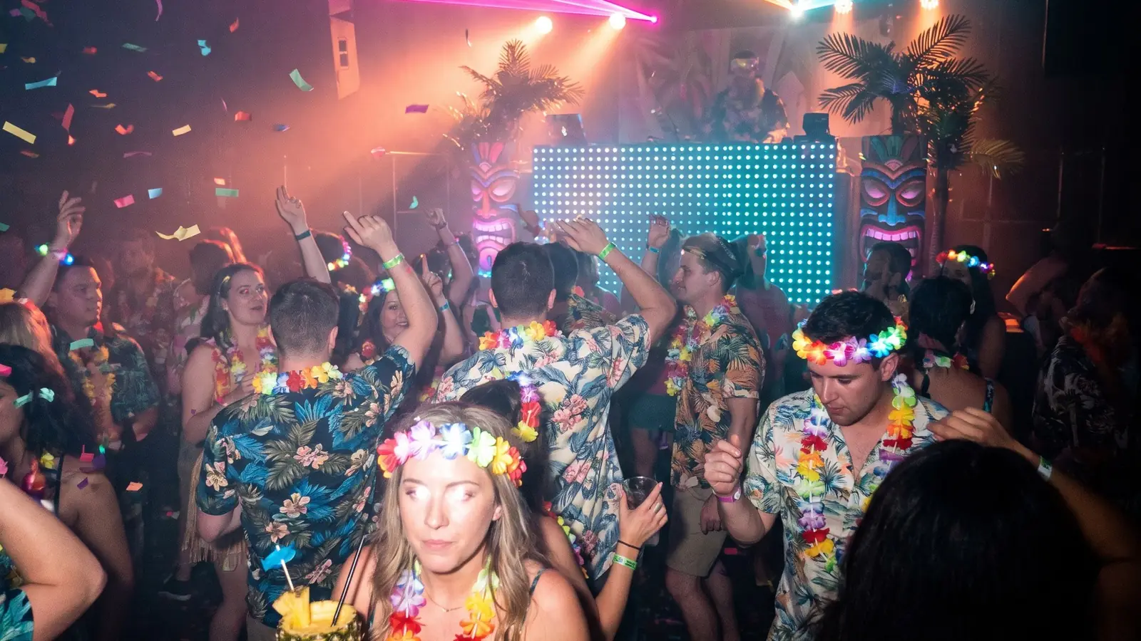 A packed dance floor of Hawaiian shirts and flower leis, with confetti and pink and orange laser lights